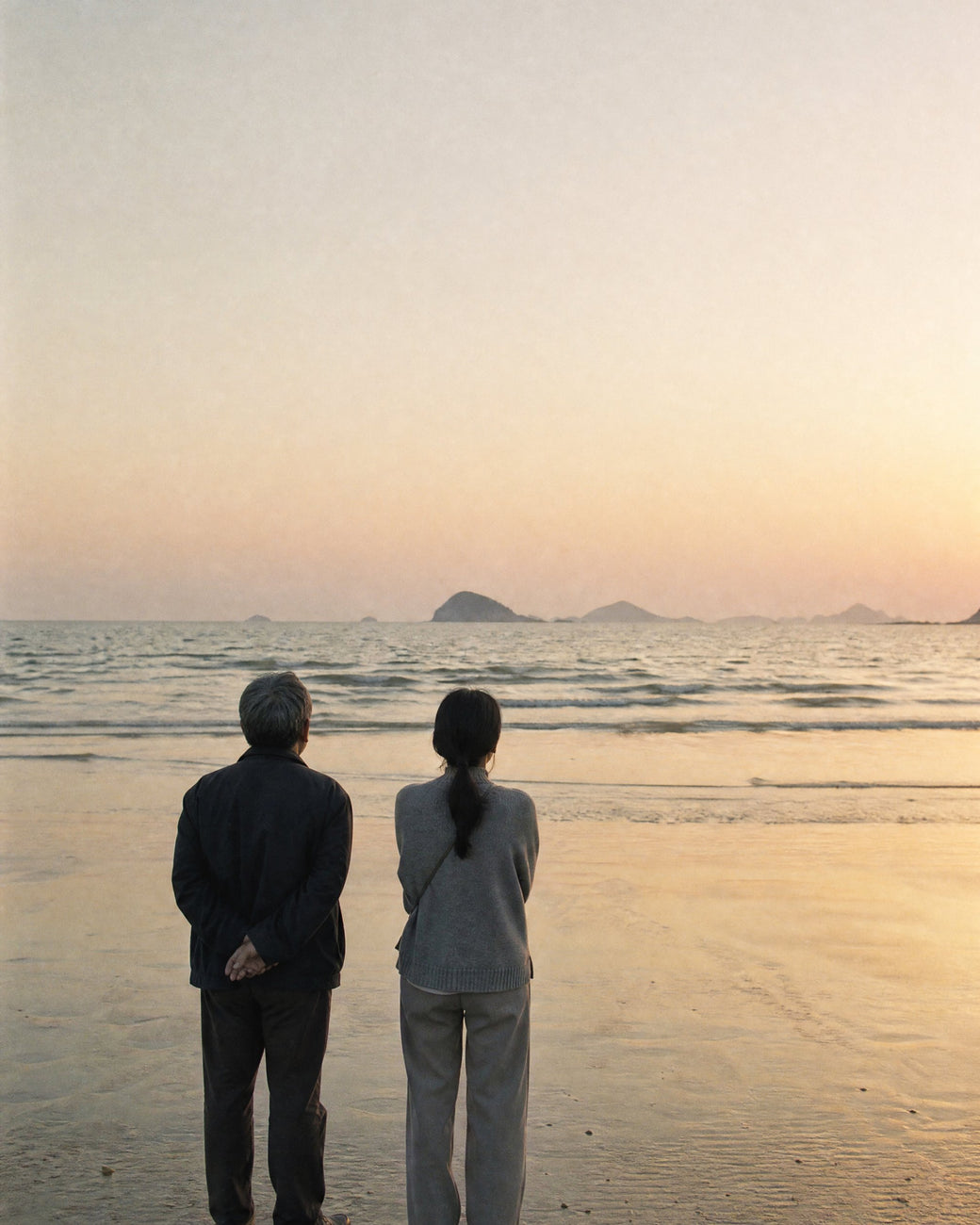 A father and his grown daughter standing together on a beach at sunset, seen from behind, looking out at the sea